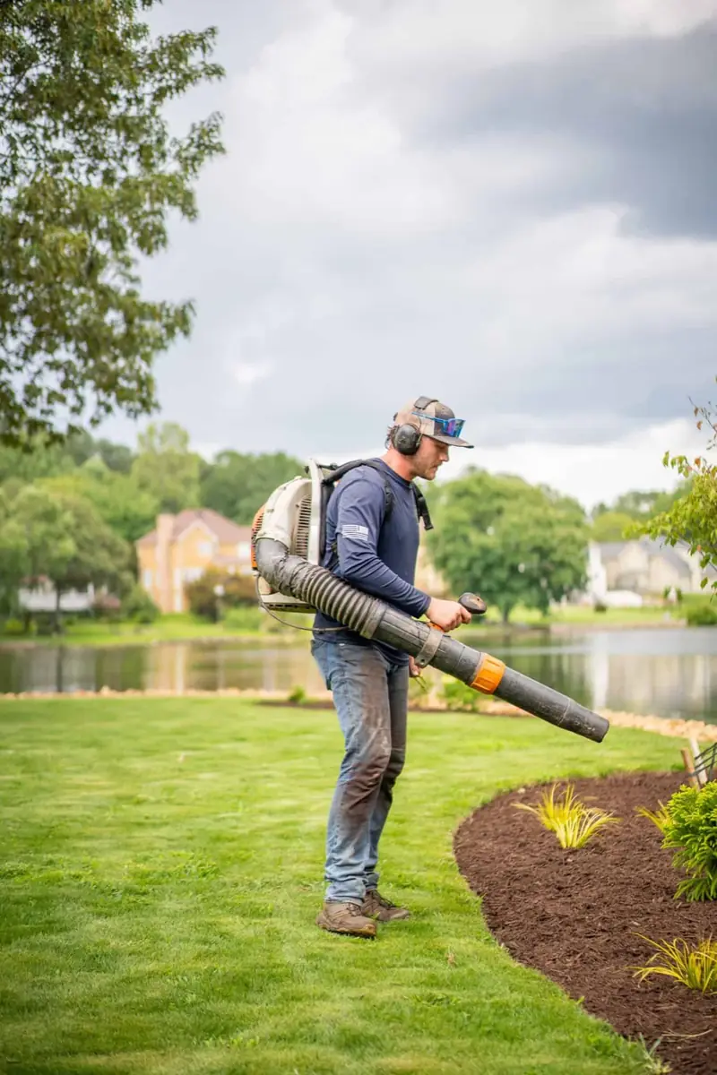 KalKare crew member using a backpack blower to clear a lakeside lawn and mulched bed