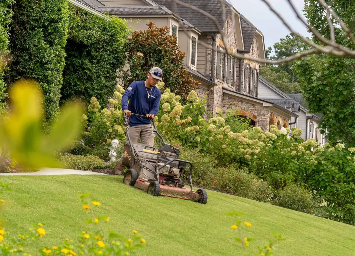 KalKare crew member mowing a sloped lawn past blooming hydrangeas at a brick home