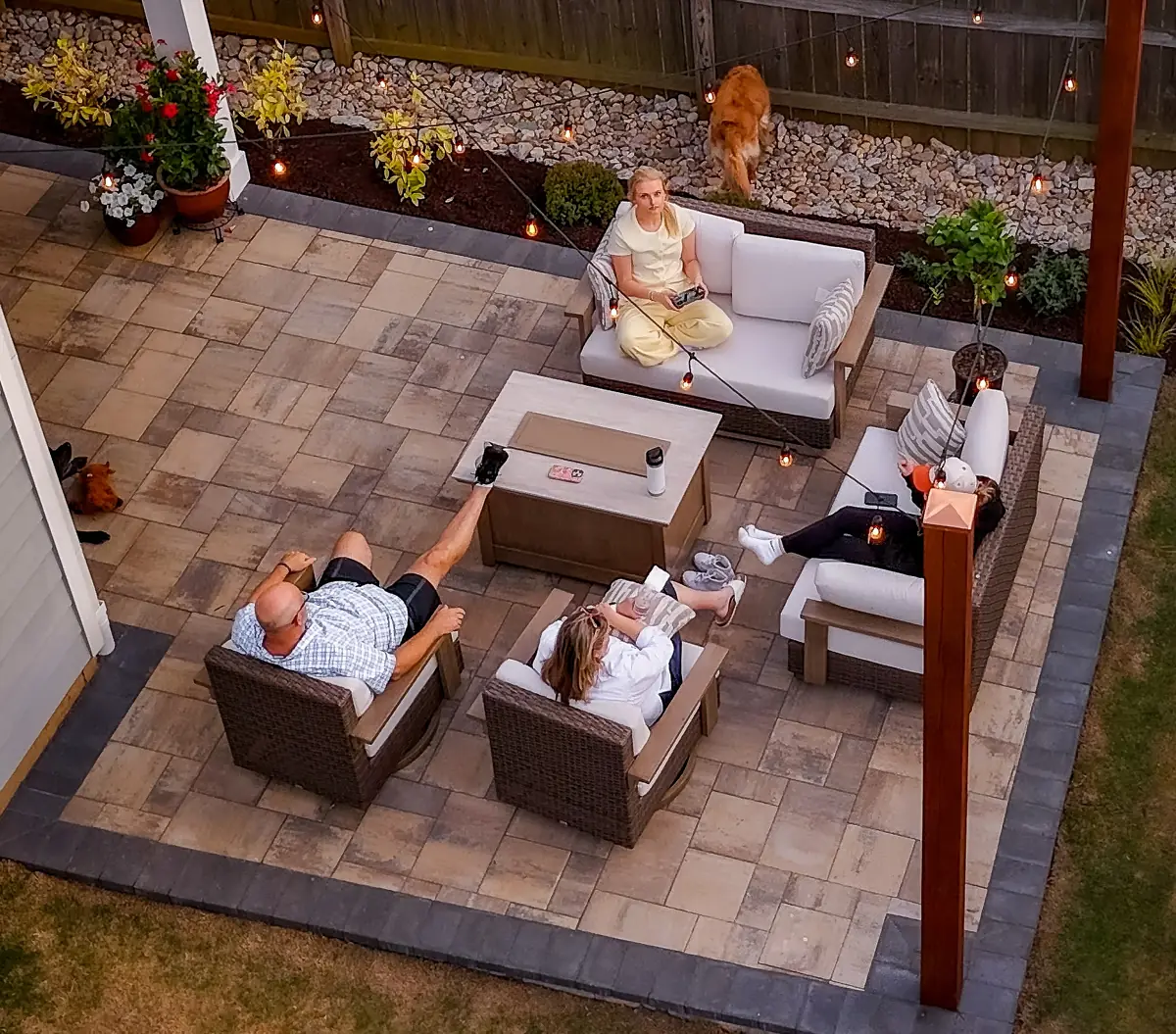 Family relaxing on a paver patio around a gas fire table under string lights at dusk