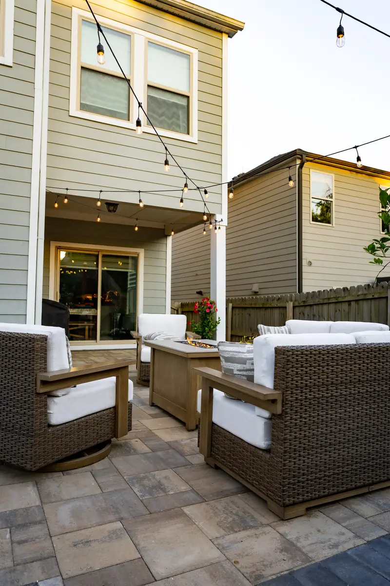 Paver patio with wicker lounge seating, a fire table and string lights beside a covered porch