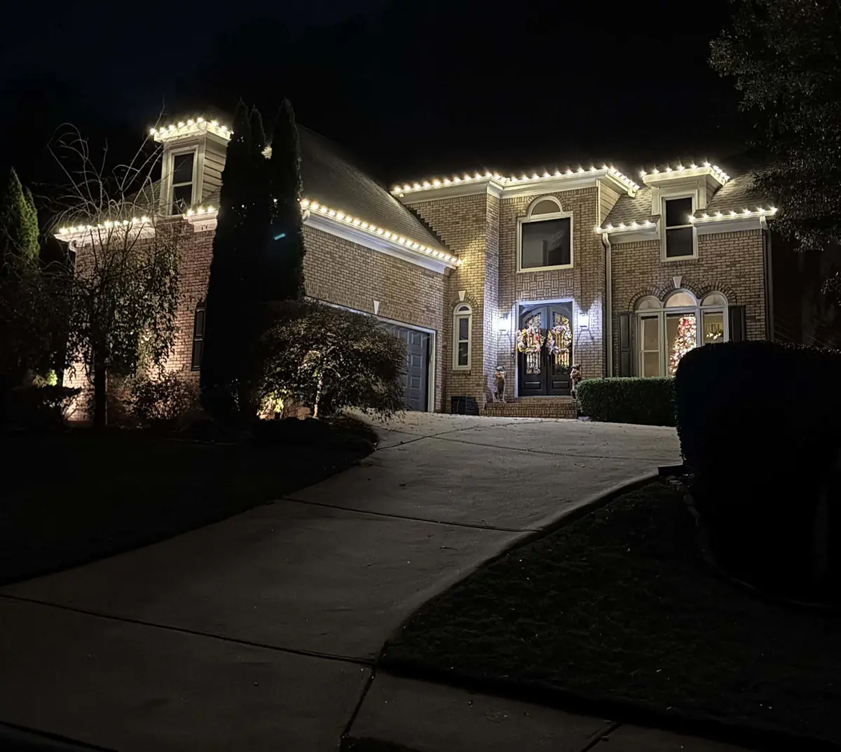 Brick home at night with warm-white roofline lighting and lit wreaths in the windows