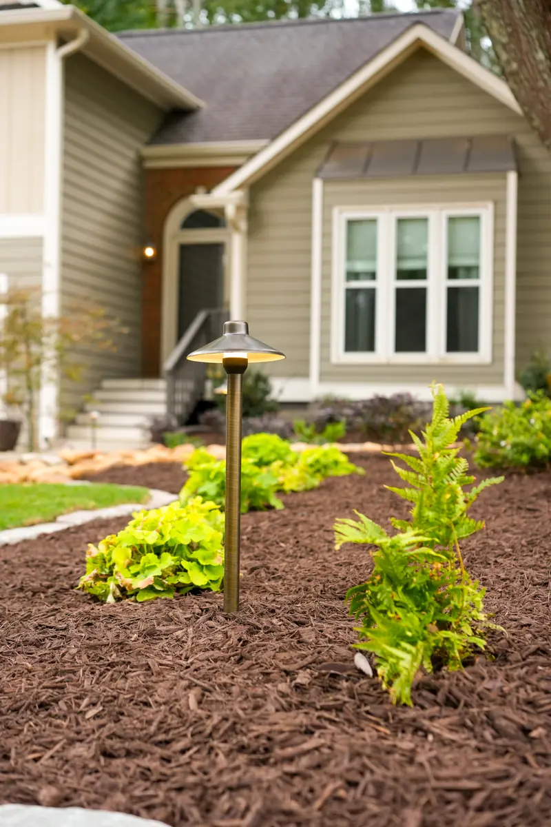 Brass path light illuminating a mulched foundation bed in front of a home