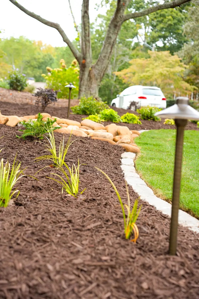 Brass path light in a stone-edged planting bed with ornamental grasses along a driveway